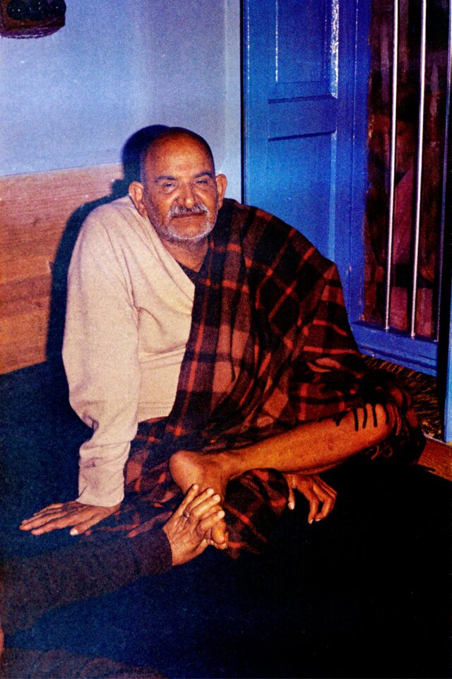 Neem Karoli Baba seated against a wall, wrapped in a plaid blanket
